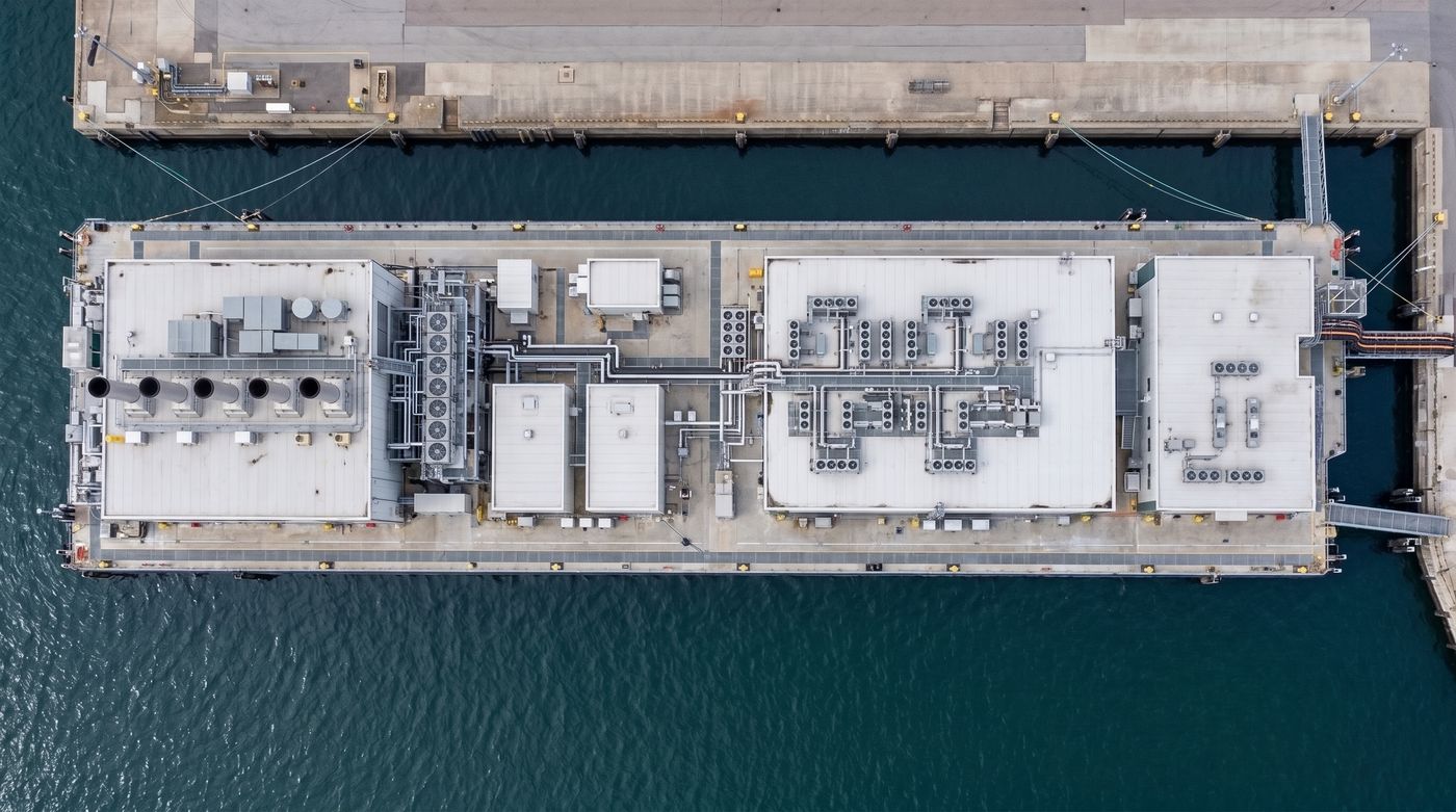 Concept render, top-down aerial view of the unit moored along a quay: power plant with exhaust stacks, electrical blocks, the data-hall roof with cooling equipment and the support block.